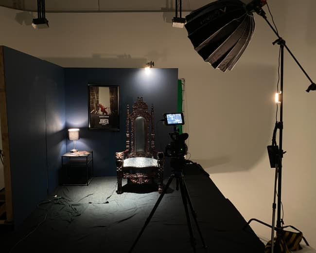 A photography studio setup with a decorative throne-like chair, a table lamp, a poster on the wall, and a camera on a tripod pointed at the chair.