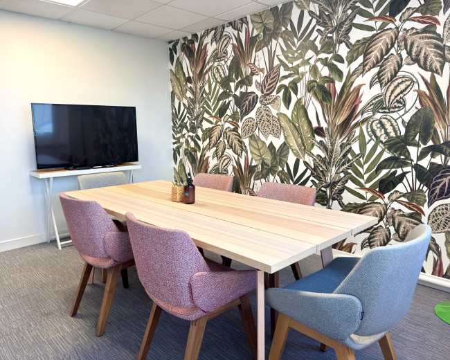 The image shows a conference room with a wooden table surrounded by colorful chairs and a large television mounted on the wall, which is adorned with a botanical wallpaper.