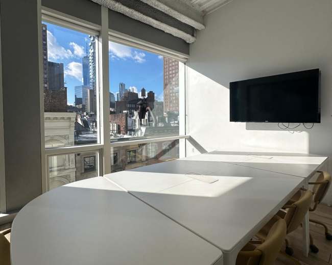 A bright meeting room features a white table and chairs, with large windows showcasing a view of city buildings and a television mounted on the wall.