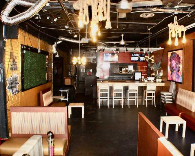 The image shows a dimly lit bar interior with seating areas, a counter with stools, and decorative elements such as hanging lights and ropes.