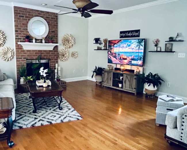 A living room features a brown brick fireplace, a large television mounted on a wall, and a combination of decorative elements and furniture including a gray sofa, a coffee table, and a patterned rug.