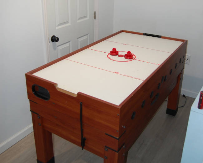 The image shows a wooden air hockey table positioned against a white wall with a closed door.