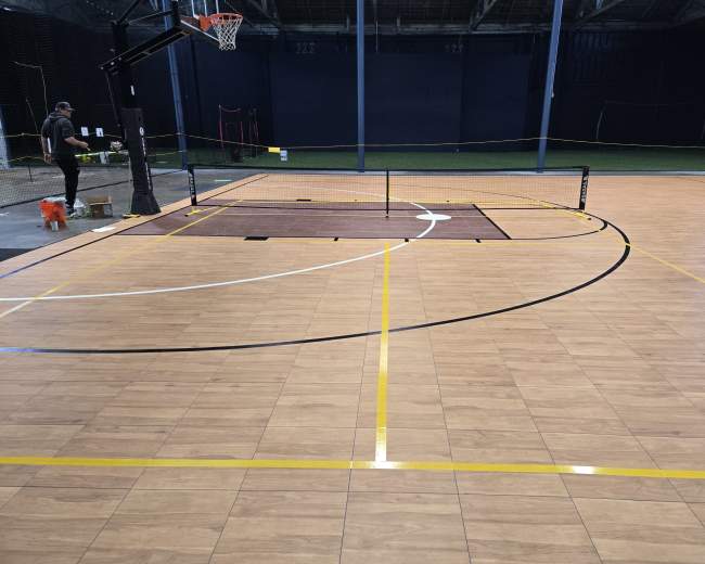 The image shows an indoor sports facility with a basketball hoop and a court featuring distinct markings for basketball and possibly other sports.