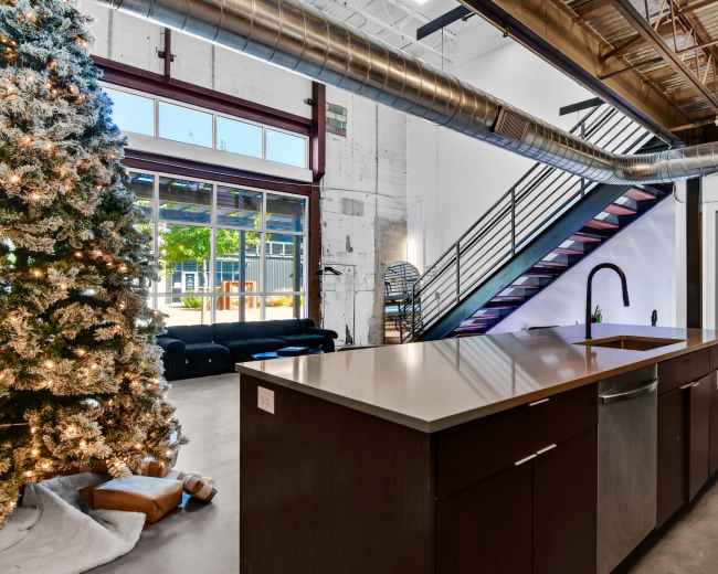 The image features a modern kitchen with a large island and a Christmas tree nearby, set in an industrial-style space with exposed beams and a staircase.