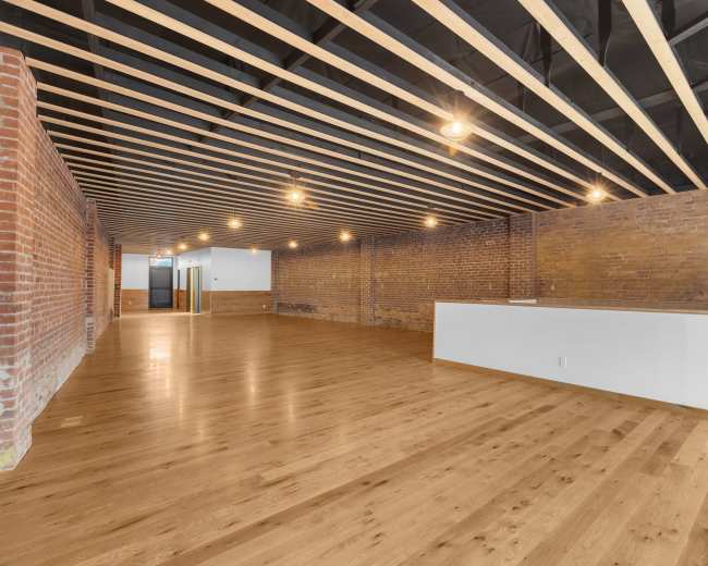 The image shows an empty commercial space with exposed brick walls, wooden flooring, and a ceiling with visible beams and lighting fixtures.