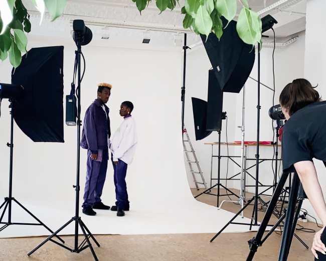 Two individuals in matching purple outfits stand in front of a white backdrop surrounded by professional photography equipment and lighting.
