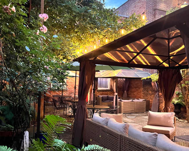 The image shows a cozy outdoor seating area adorned with string lights, surrounded by lush greenery and flowers.