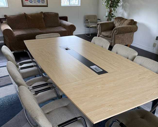 The image shows a large wooden conference table surrounded by eight chairs in a brightly lit room with a brown couch and a potted plant in the background.