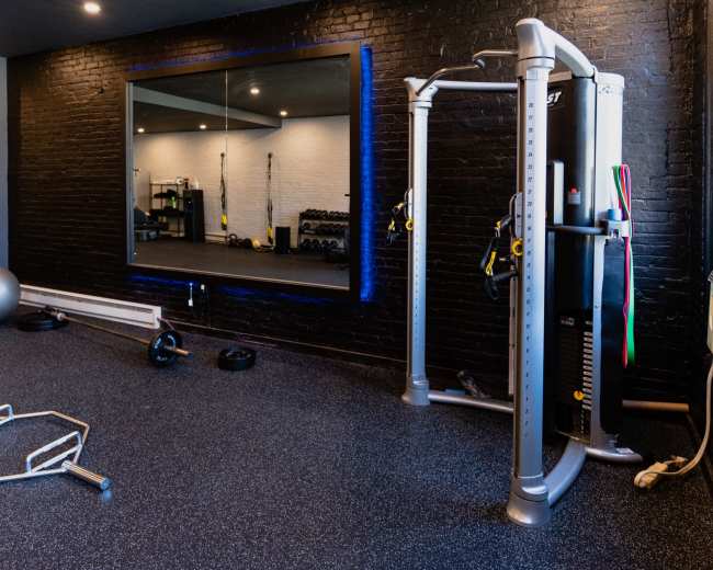 A modern gym interior featuring a cable machine, free weights, and a large mirror against black brick walls.