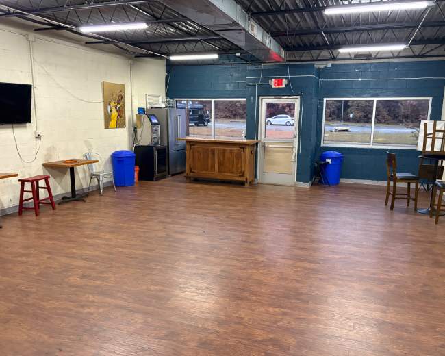 The image depicts an empty indoor space with wooden flooring, featuring a bar area, several chairs and tables, a television, and blue trash bins against the walls.
