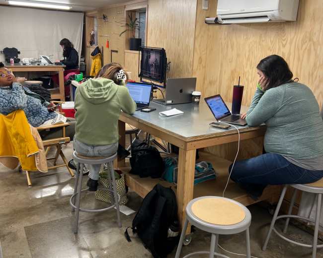 Two individuals work on laptops at a shared table, while another person is seated in the background of a casual workspace.