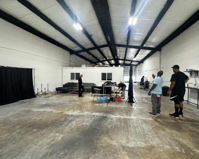 A spacious, partially empty warehouse features a few people setting up equipment, with black curtains and a sofa visible in the background.