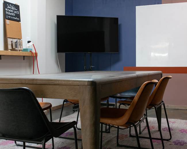 The image shows a modern conference room featuring a long wooden table surrounded by black and brown chairs, a large television mounted on the wall, and a whiteboard.