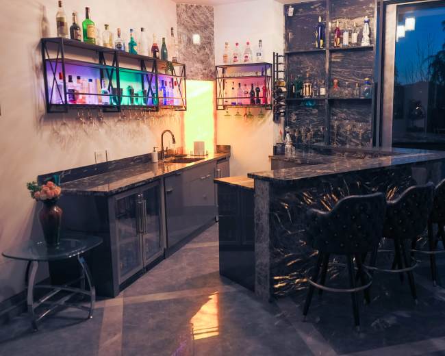 The image shows a modern home bar with dark granite countertops, illuminated shelves displaying various bottles, and seating along the bar area.