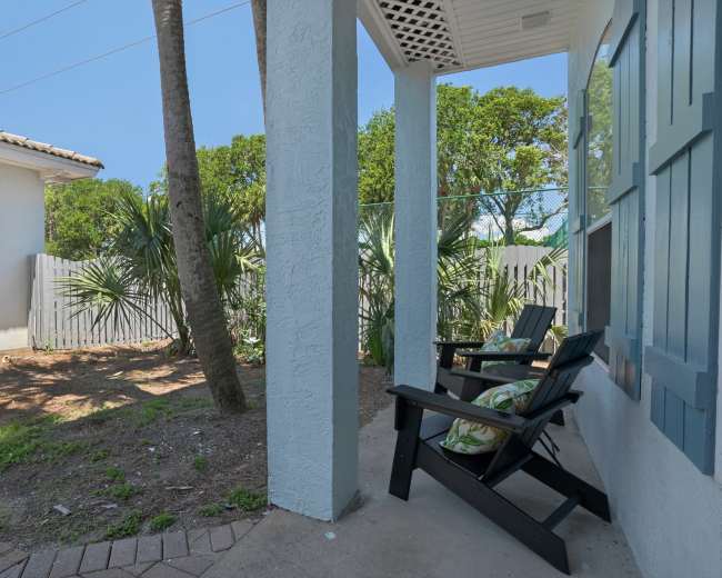 A two-chair porch area looks out onto a backyard with greenery and a white fence adjacent to a residential home.