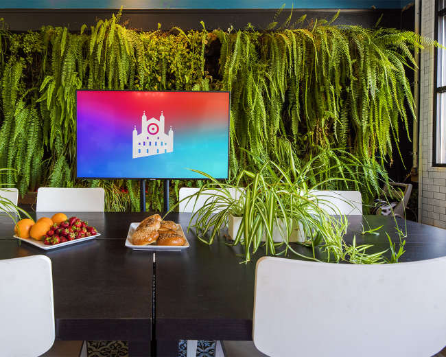 A dining table is set with fruit and pastries in front of a large screen displaying a colorful logo, surrounded by lush greenery on the walls.