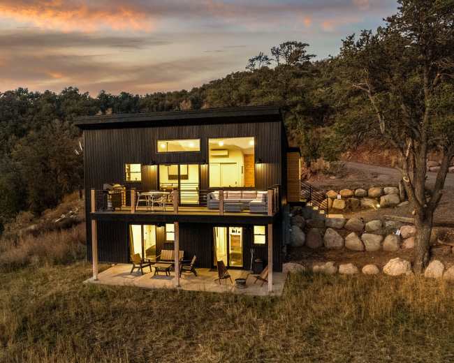 A modern black house with large windows is positioned on a hillside, surrounded by grass and boulders, as the sun sets in the background.