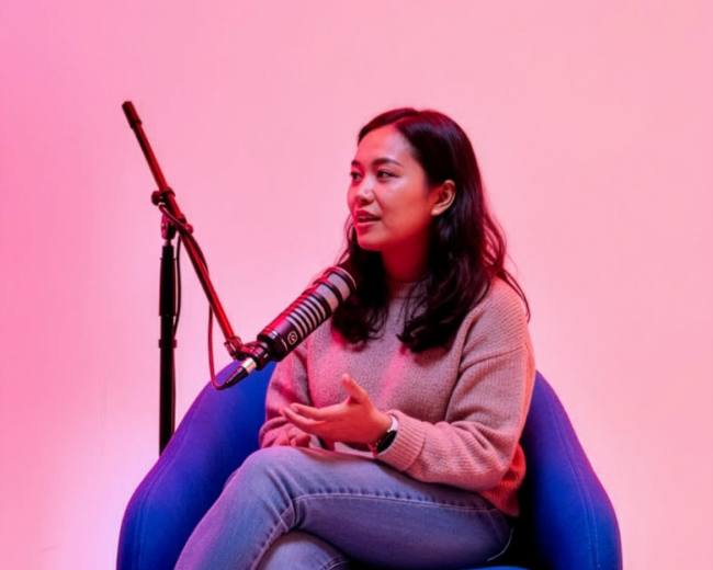 A woman sits on a blue chair, speaking into a microphone against a pink and purple backdrop.