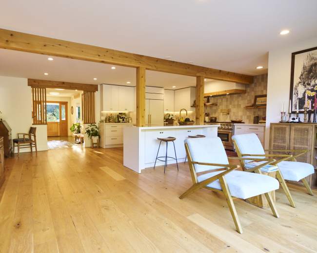 The image shows a modern open-concept living space featuring wooden beams, light-colored cabinetry, and two armchairs facing a kitchen area.