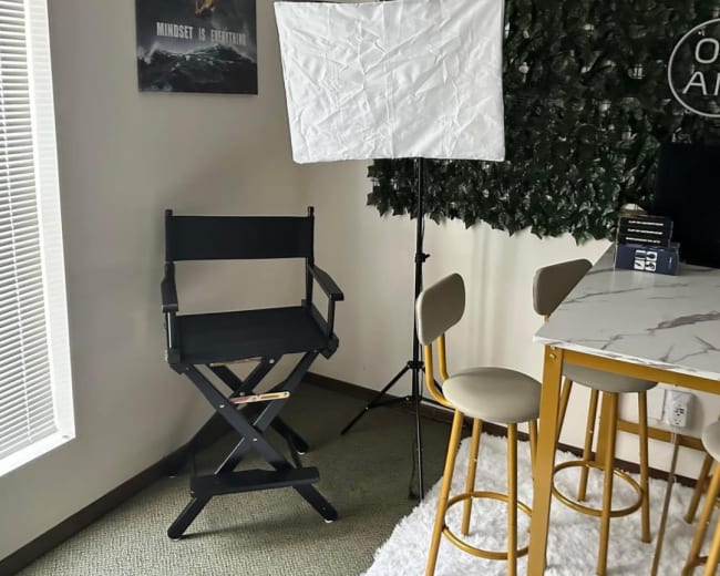 A corner of a room with a director's chair, a softbox lighting setup, and a small bar table with chairs beside a wall covered in greenery.