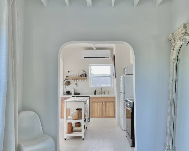 A bright, minimalistic kitchen features wooden cabinets and a stainless steel stove, visible through an archway in a white-walled room.