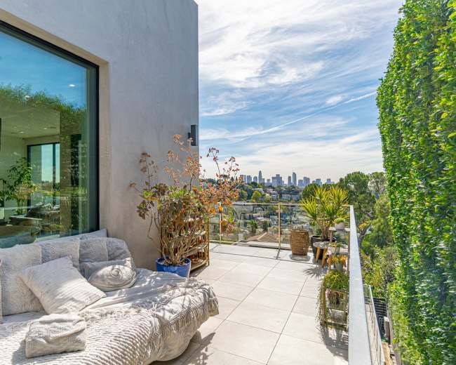 A modern outdoor terrace with a plush seating area, potted plants, and a view of a city skyline under a blue sky.