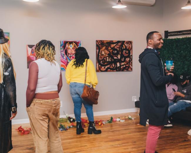 A group of people stands in an art gallery, viewing colorful paintings on the walls while some engage in conversation.