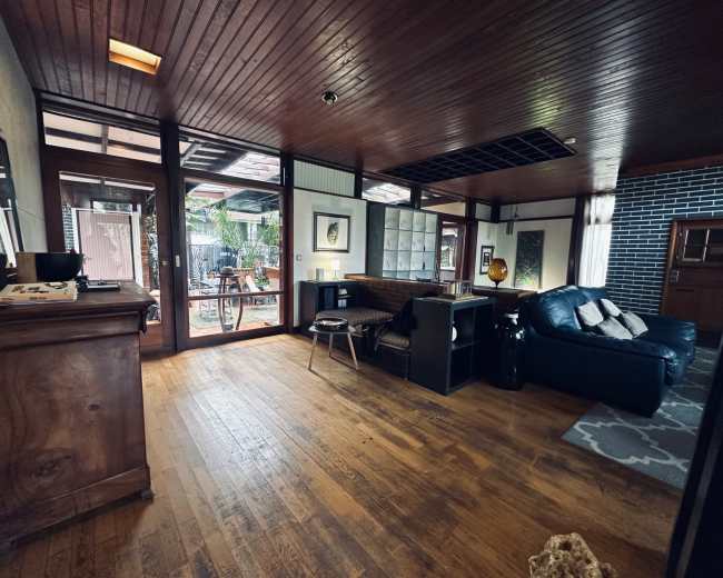 The image shows a spacious interior of a home with wooden flooring, large windows, and a seating area that leads to an outdoor space.