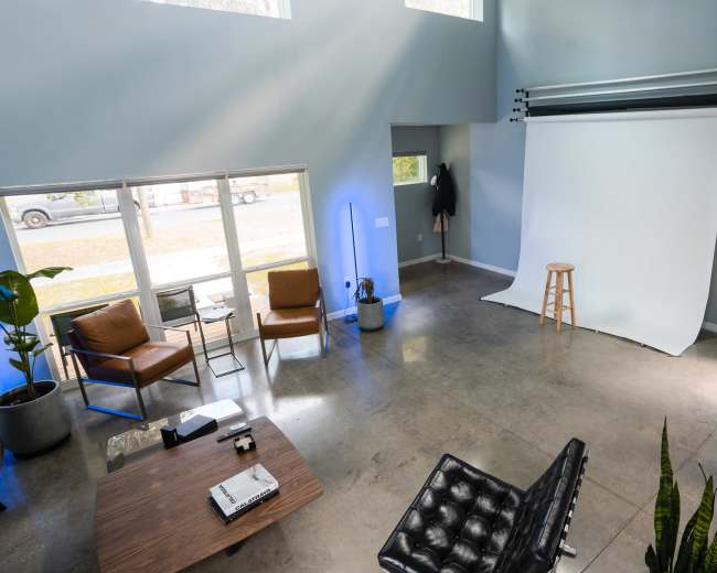 A spacious studio with a white backdrop, a wooden stool, and modern furniture, illuminated by natural light from large windows.