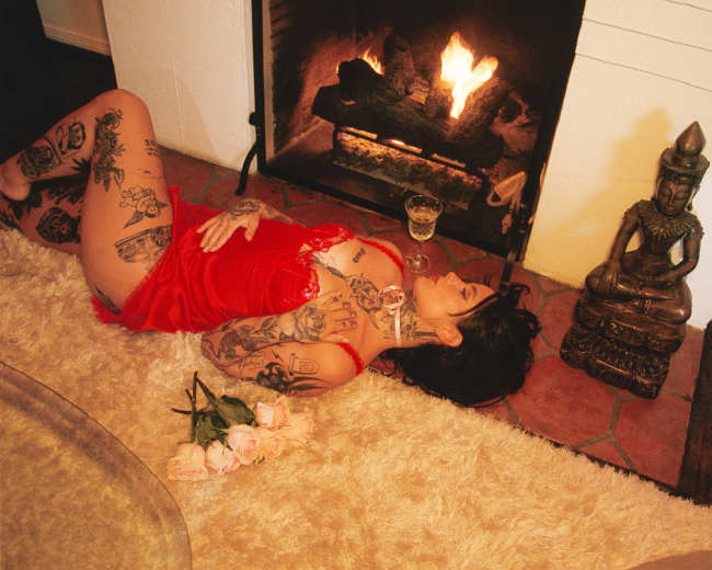 A person lies on a soft rug beside a fireplace, holding a cocktail, with a statue and a bouquet of roses nearby.