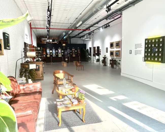 The image shows an art gallery featuring a spacious layout with various framed artworks on the walls, some seating in the foreground, and plants placed throughout the space.
