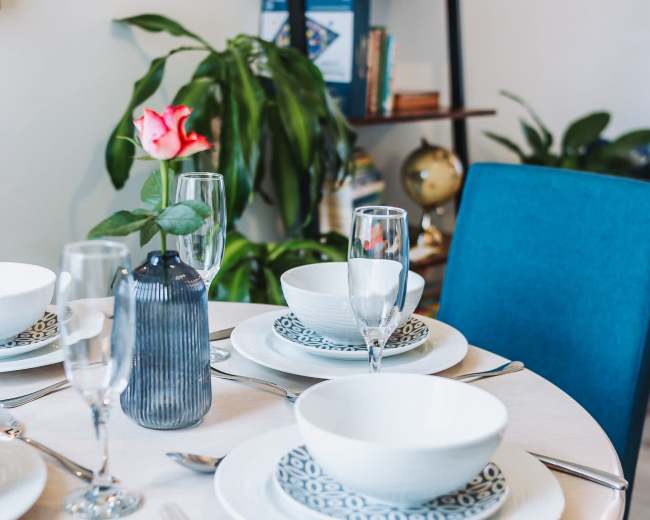 A round dining table is set with white bowls, decorative plates, and glassware, accompanied by a vibrant rose in a vase, against a backdrop of a bookshelf and indoor plants.