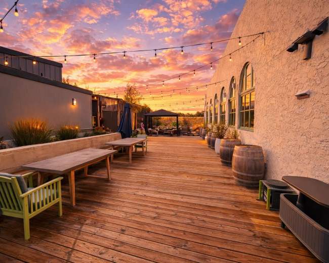 The image shows a wooden deck with tables and chairs, surrounded by potted plants, under a colorful sunset sky illuminated by string lights.