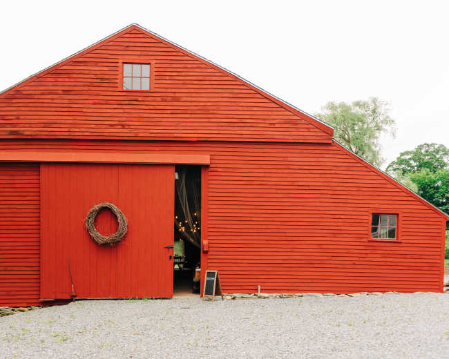 A large red barn with a door featuring a wreath is situated on a gravel path.