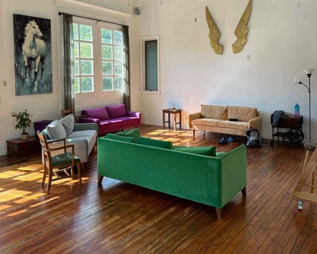The image shows a spacious room with wooden floors, large windows, and a mix of seating options, including a green sofa and pink chairs.