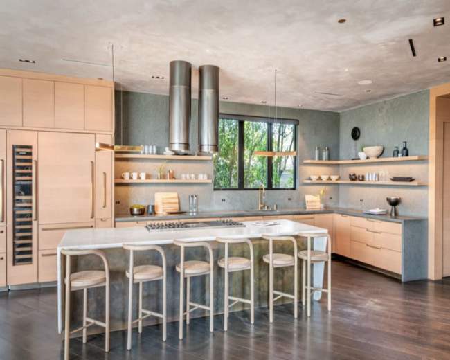 The image shows a modern kitchen with light wood cabinetry, a large central island, and stainless steel appliances, featuring a large window that allows natural light to enter.
