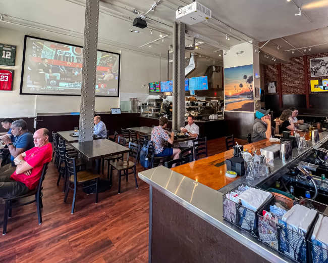 The image shows the interior of a restaurant with tables and chairs filled with patrons, large screens displaying sports on the walls, and a counter area with various items organized.