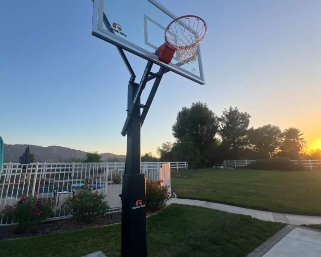 A basketball hoop stands on a driveway at sunset, with a landscaped yard and mountains in the background.