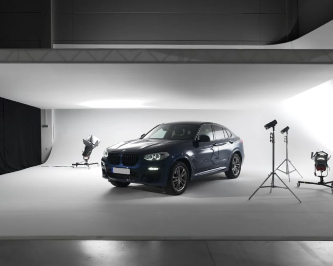 A dark blue BMW is positioned on a white studio backdrop, illuminated by various lighting equipment.