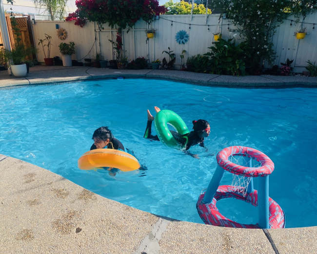 Two children are playing in a swimming pool, one holding an orange float and the other using a green float, with a basketball hoop positioned at the pool's edge.