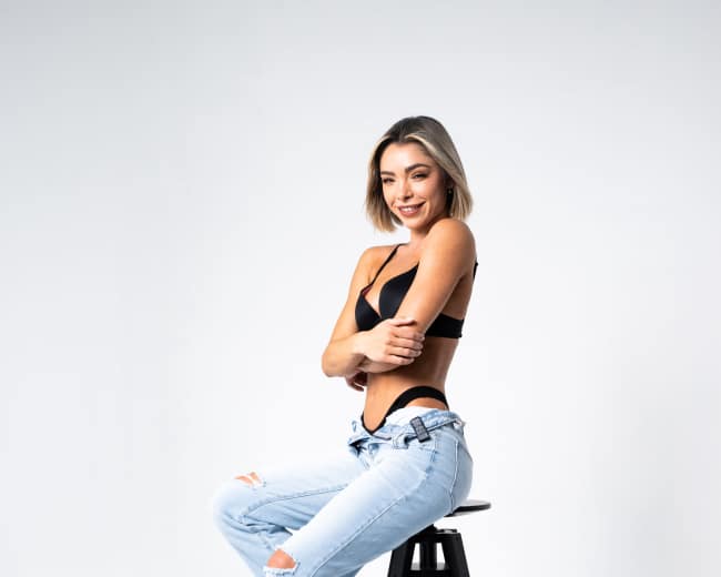 A woman in a black top and ripped light-wash jeans sits on a black stool against a plain background.