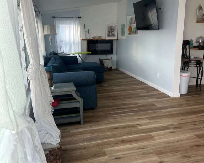 The image shows a living room with blue sofas, light wood flooring, and a mounted television on the wall.