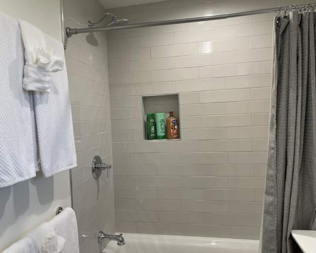 The image shows a clean shower area with a tiled wall, a small niche for toiletries, and hanging towels.