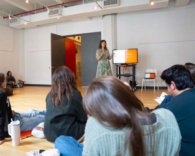 A woman stands in front of an audience seated on the floor in a bright, open room, delivering a presentation.