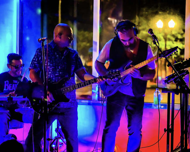 A band performs on stage with a drummer, guitarist, and singer under colorful lighting.