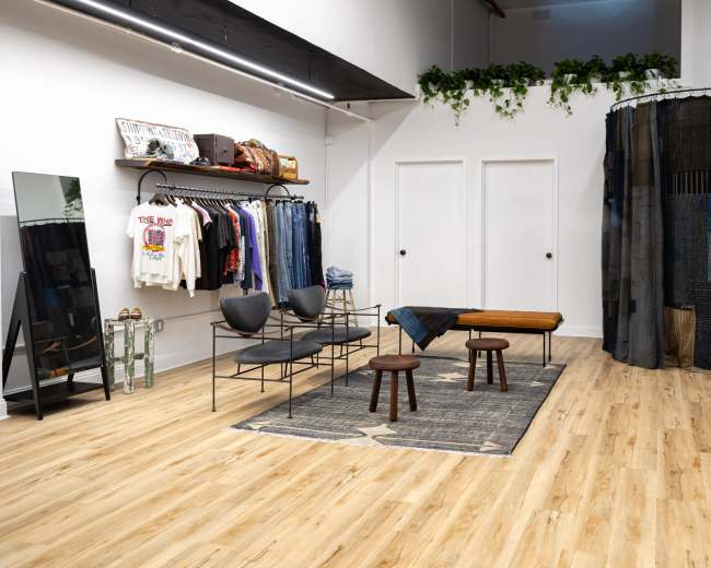 The image shows a modern clothing store interior with racks of denim and apparel, a mirror, a wooden bench, and a patterned rug.