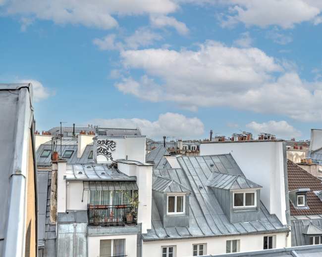 A view of rooftops in a city, featuring a mix of metal and tiled roofs, balconies, and scattered graffiti under a partly cloudy sky.