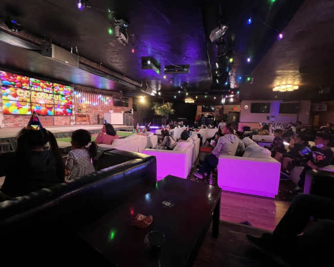 A lively lounge area with white couches, colorful lighting, and a large screen displaying the word "COLORFUL" while guests gather and socialize.