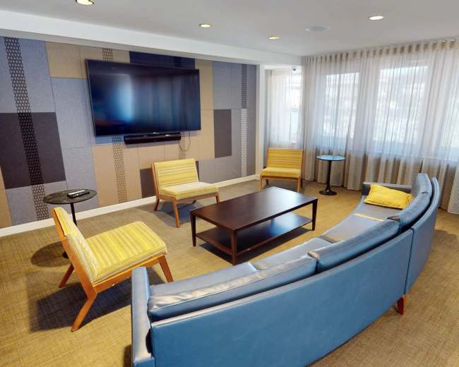 The image shows a modern living room with a large television mounted on the wall, a circular blue couch, two yellow-striped armchairs, and a coffee table in the center, surrounded by floor-to-ceiling windows with sheer curtains.