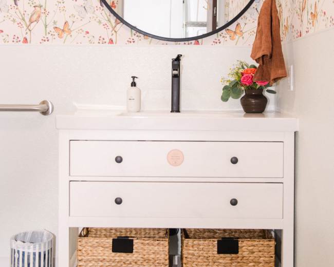 A bathroom with a round mirror above a white vanity featuring two drawers and woven baskets below, set against a colorful bird-themed wallpaper.